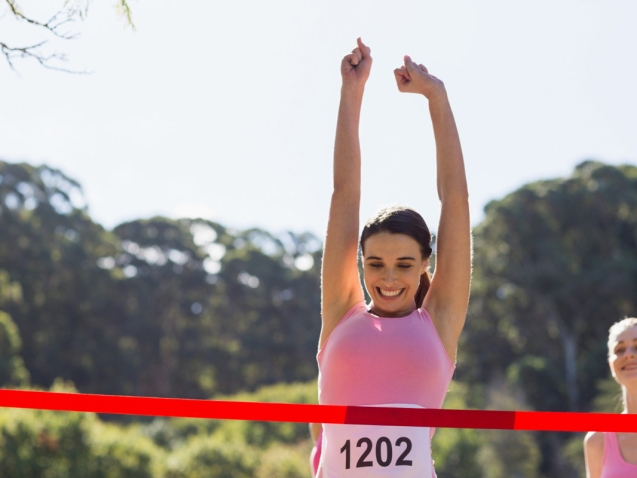 Mulher comemora erguendo os braços ao cruzar a linha de chegada de uma corrida de rua